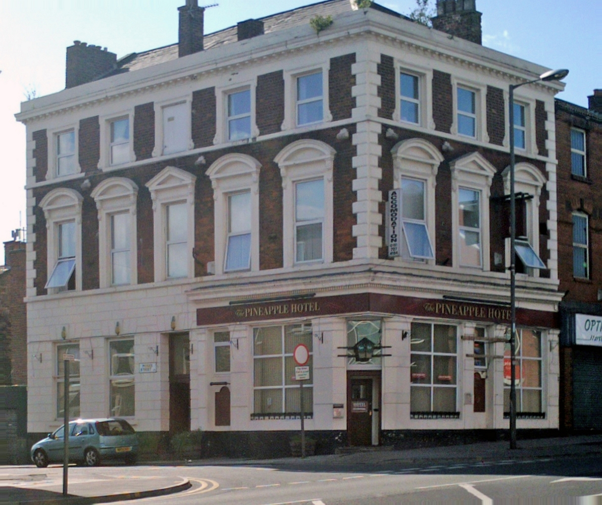 Picture of a hotel on a corner street. Three story brick and white building called the PINEAPPLE HOTEL. A car sits out front.