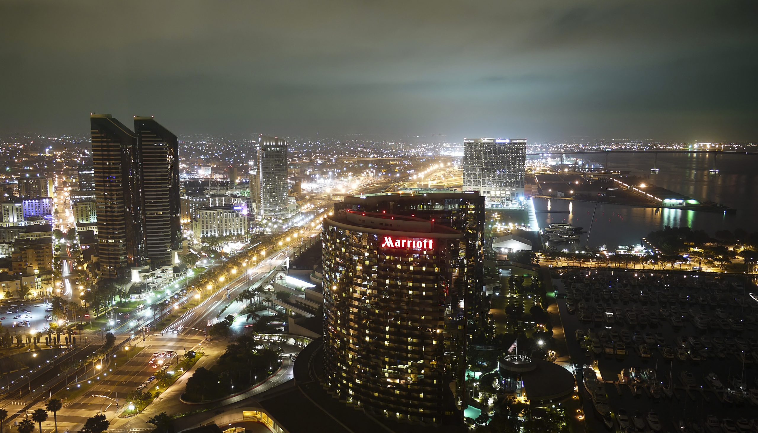 A photograph of the San Diego cityscape at night, lit up with lights. The San Diego Marriott in the foreground, a large, arch shaped skyscraper. Water is to the right of the Marriott, with lighted docks extending into the water. A highway is to the left, lined with streetlights. Other buildings are in the background, including two other skyscrapers to the left and two in the distance.