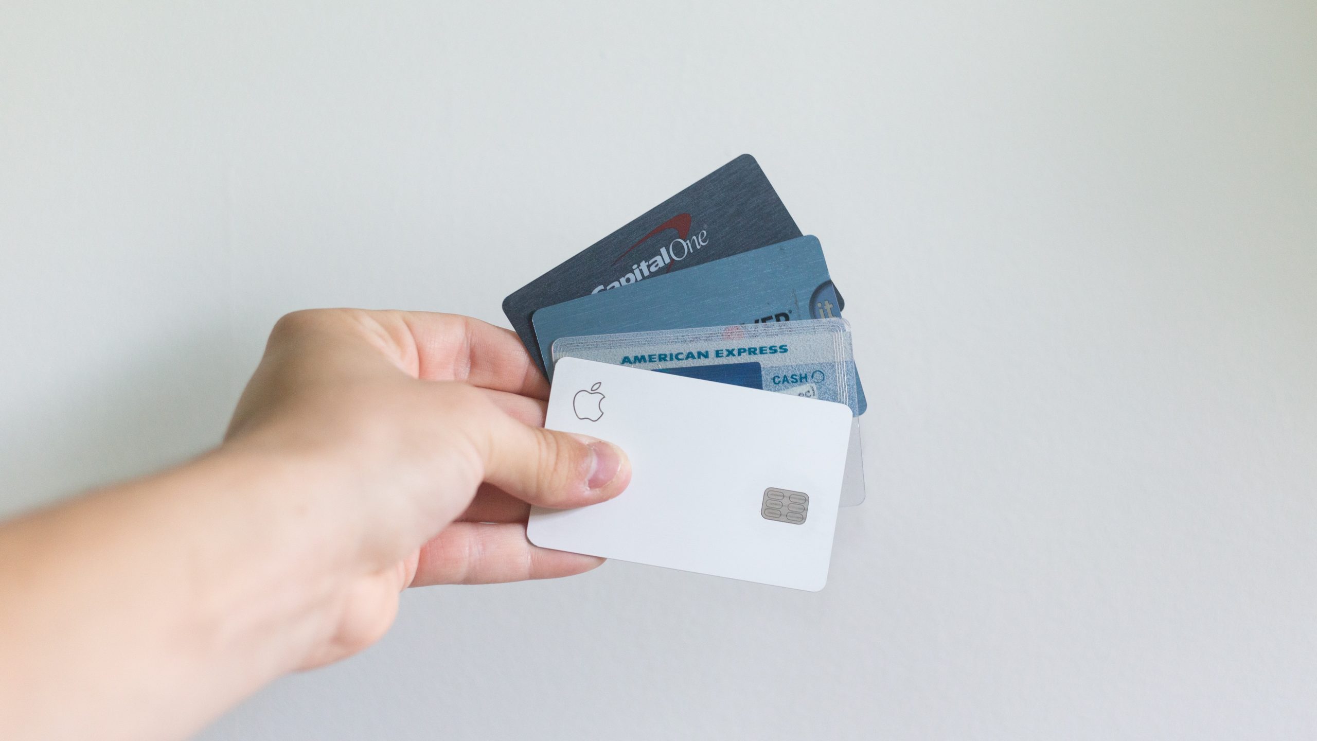 Picture of a hand holding 4 credit cards fanned out in their hand. The cards are Capital One, Discover, American Express, and Apple. The background is white.