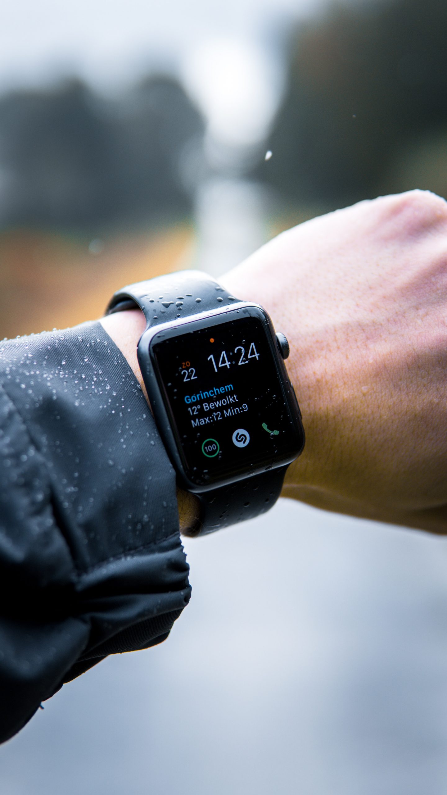 Picture of a man's wrist that has a black apple watch. The background is a road with trees on the side. It is raining.