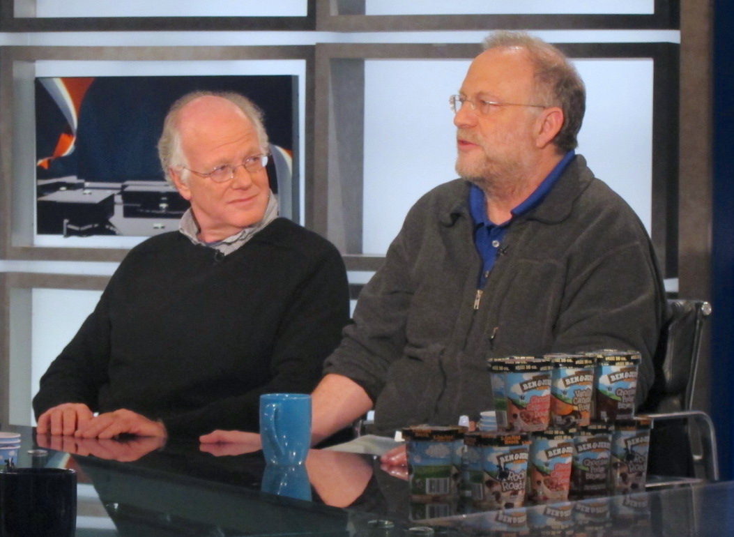 Ben and Jerry sit at a table and are talking. Both are in dark gray sweaters. Icecream pints are on the table in front of them, as well as a blue coffee mug.
