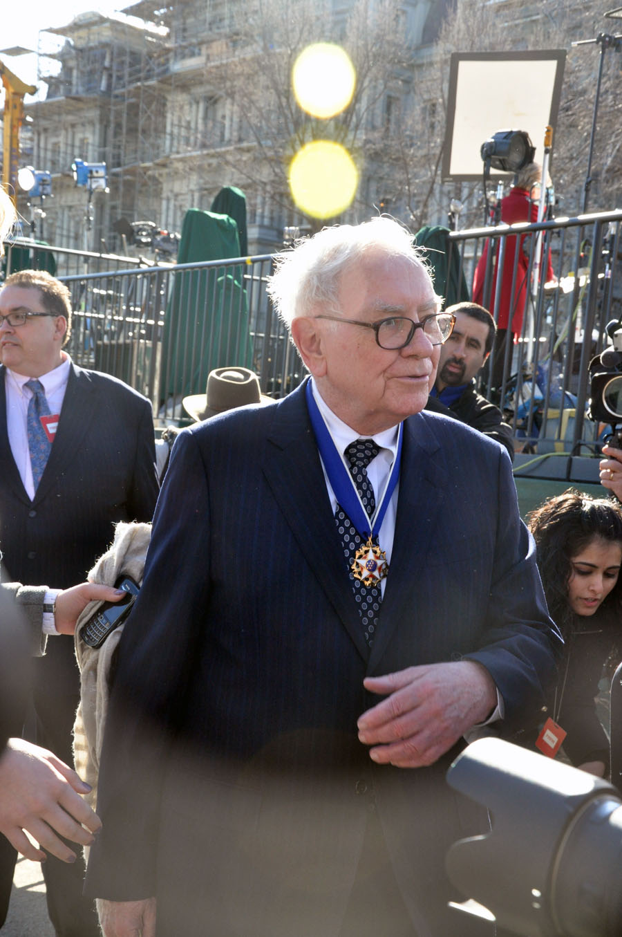 A photograph of Warren Buffet, dressed in a blue suit jacket, white shirt, and dotted tie, with his hand in front of his torso. His Presidential Medal of Freedom is around his neck. Behind him a grey brick building and a two people are visible.
