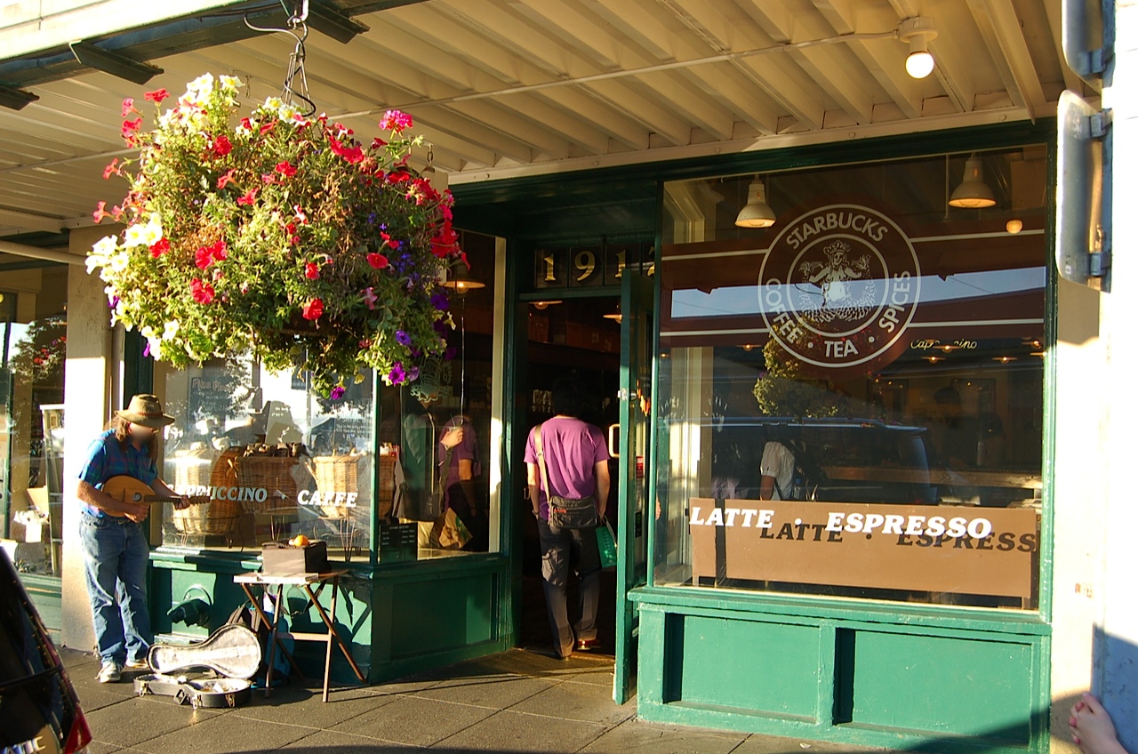 Photo of the original Starbucks. Outside storefront has green paint, bay windows on either side, and flowers hanging out front.
