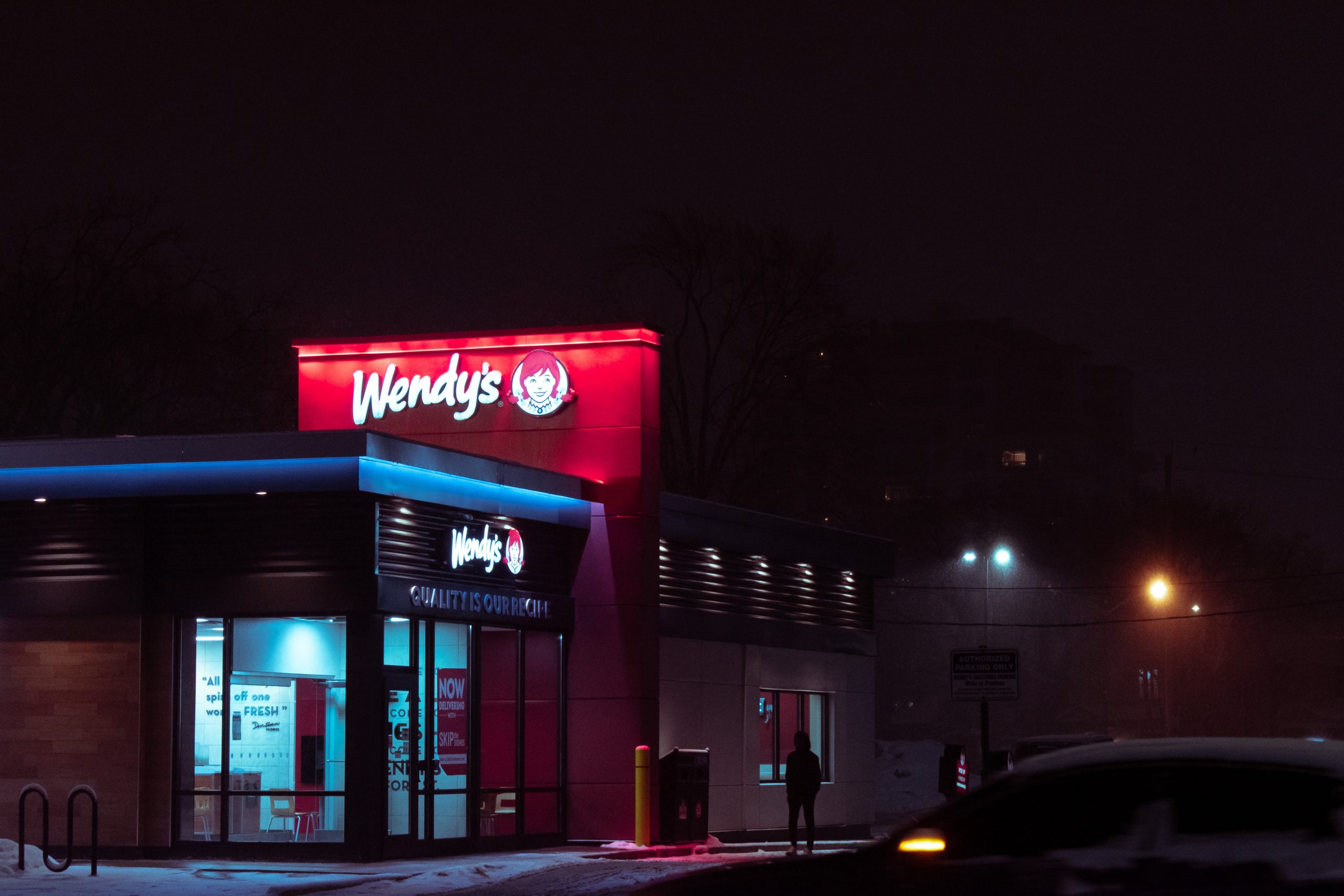 A photograph of a Wendy’s Restaurant at night, rectangular building with a flat roof, viewed from the road.