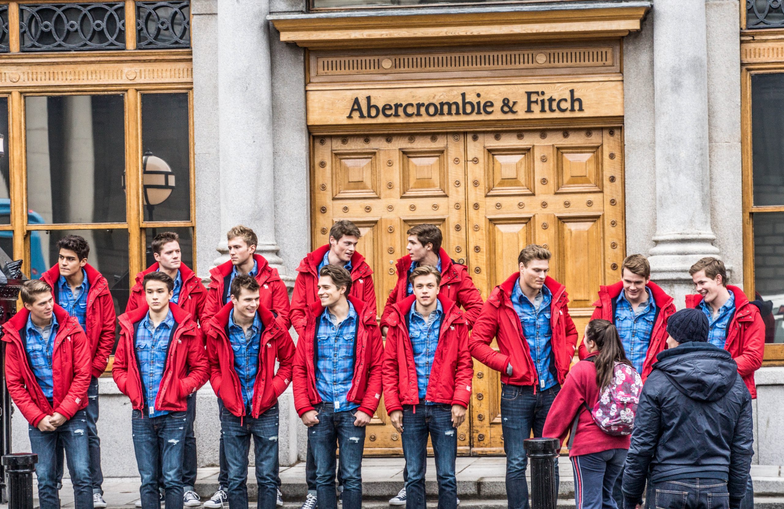 Two lines of 13 white male models, wearing the same outfit: jeans, a blue button down shirt, and a red sweater. They are standing in front of the doors of an Abercrombie and Fitch store.
