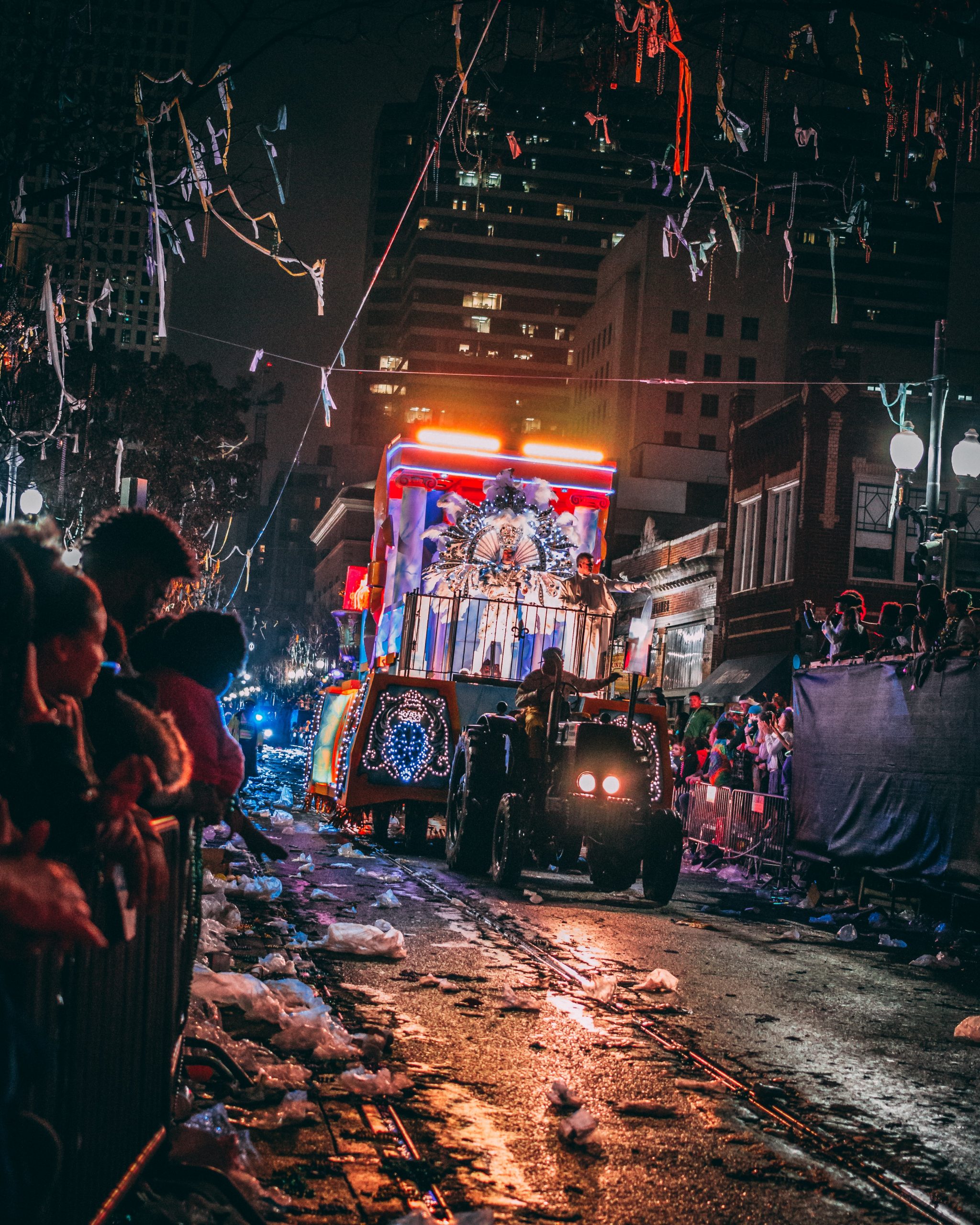 Photo at night of people lining a street as a float drives down. Streamers, confetti, and lights are strewn around the float.