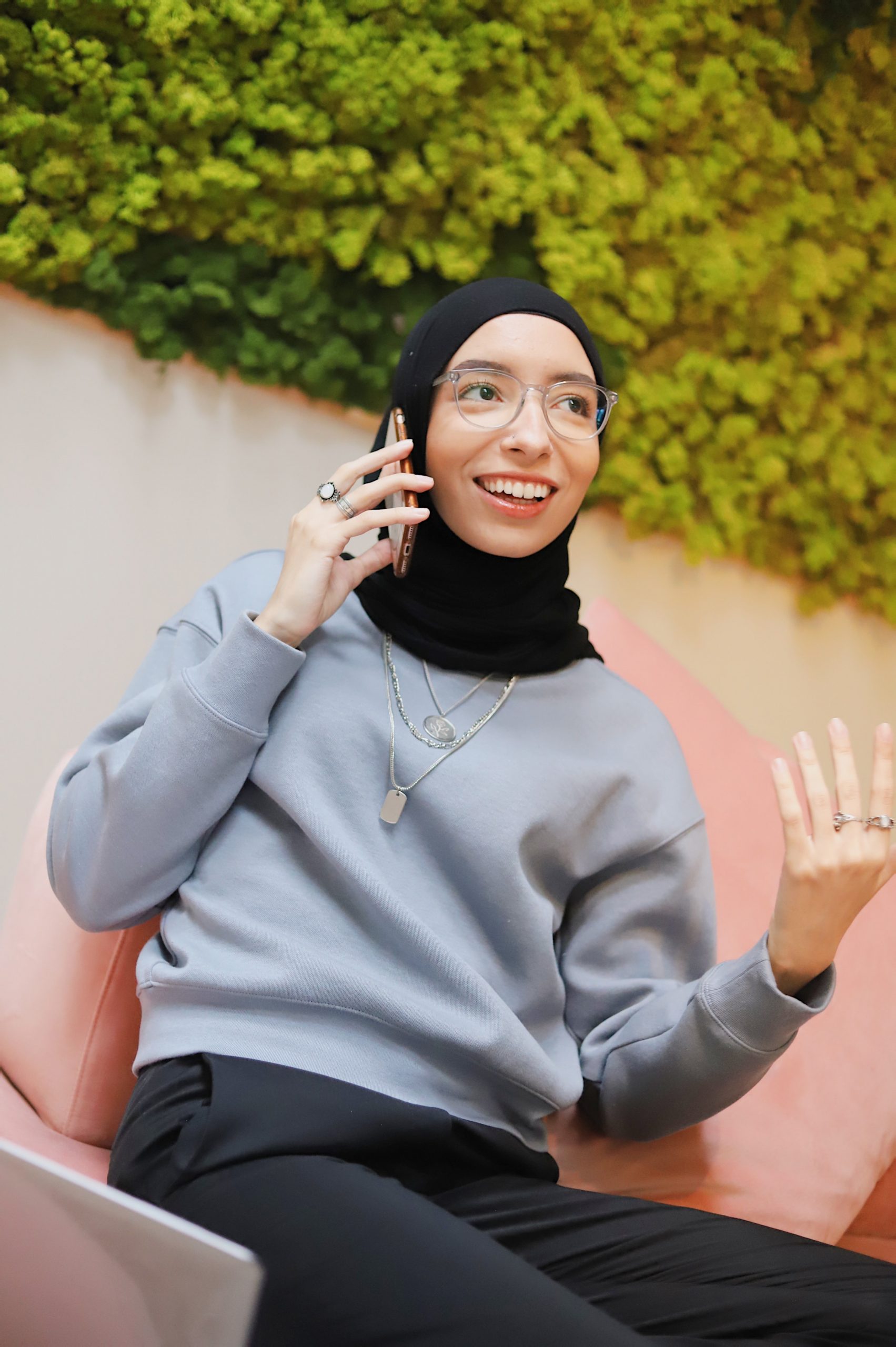 Woman wearing a black hijab and a gray sweatshirt sits in a pink chair talking on the phone.
