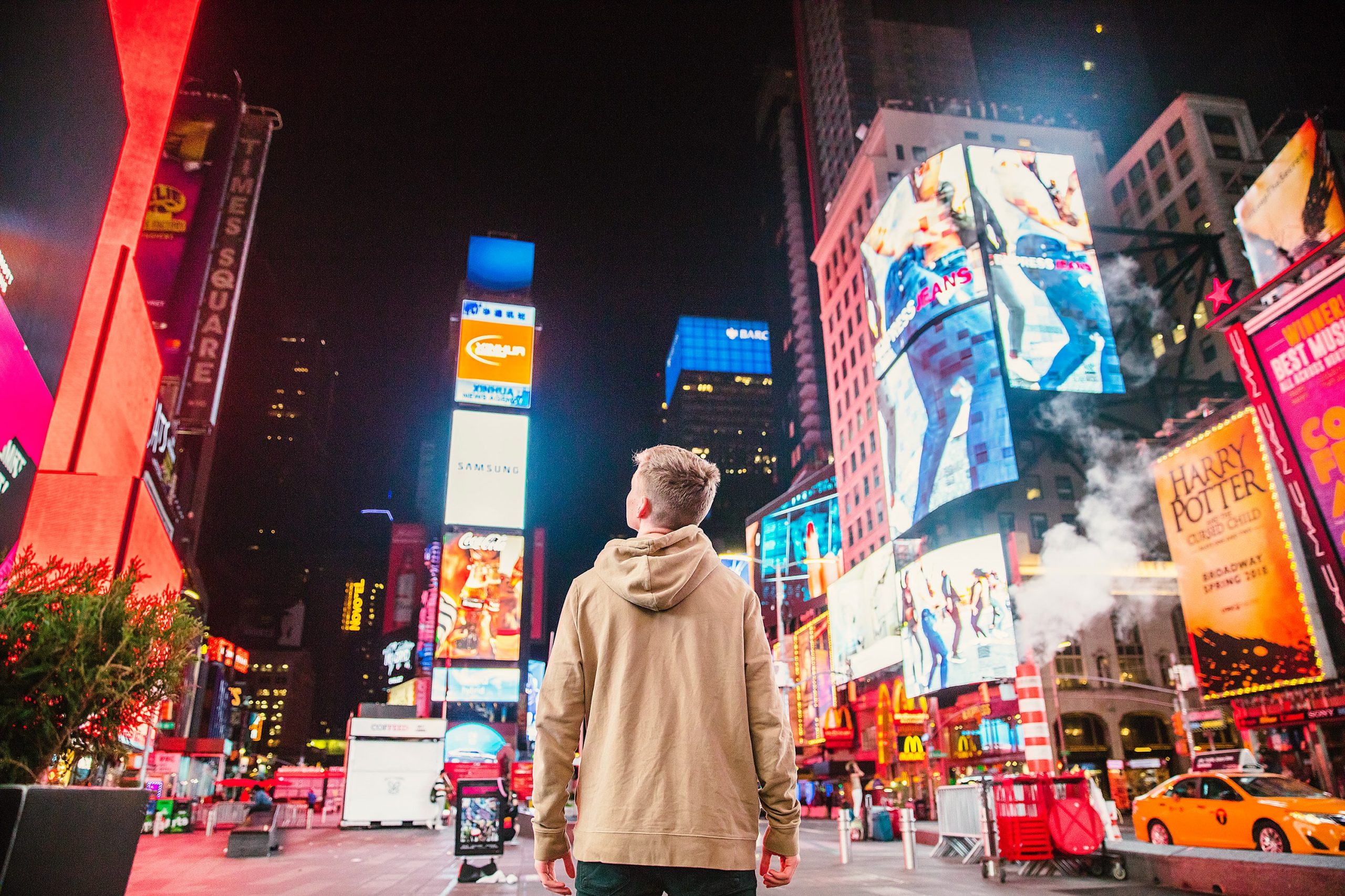 Photograph of a man standing in New York City facing many digital advertisements lit up on the sides of buildings.