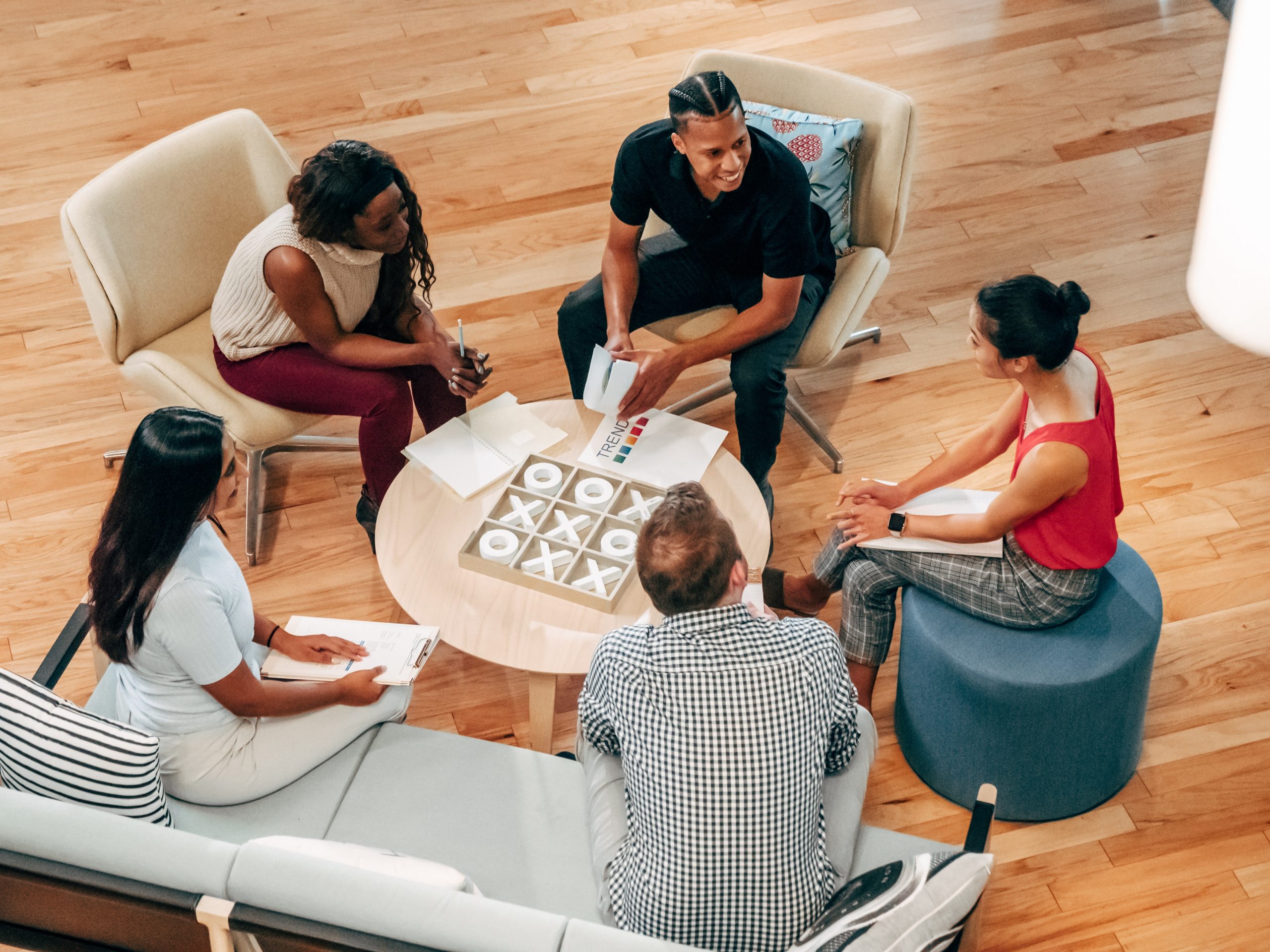 5 diverse (men/women, Black/Brown/white) people sit around a table talking and playing tic-tac-toe.