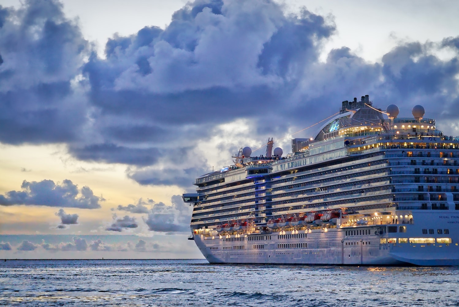 A picture of a cruise ship in the ocean at dusk. The lights on the ship are illuminated and the sky is cloudy.