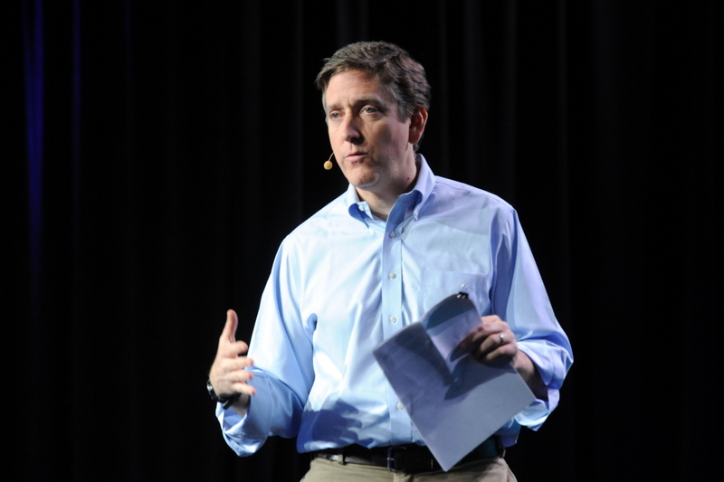 Ben Fried in a blue button up shirt, on stage, holding a pamphlet