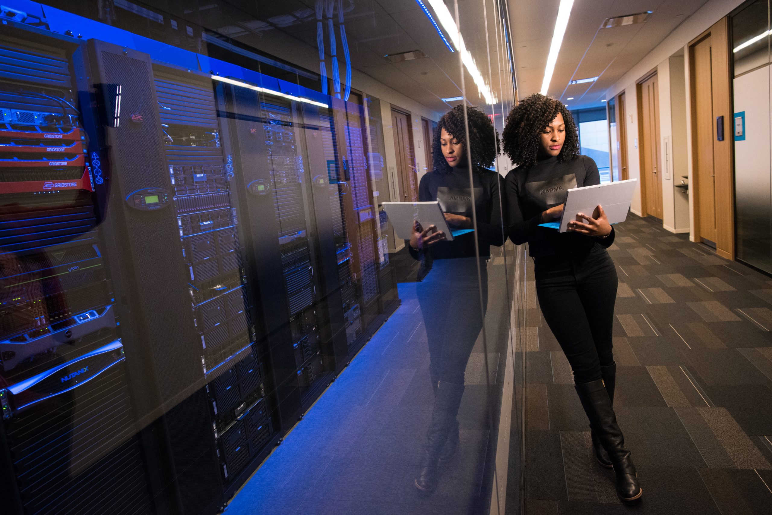 Photo of a woman holding a laptop standing in front of a room housing many computers and other technology pieces.