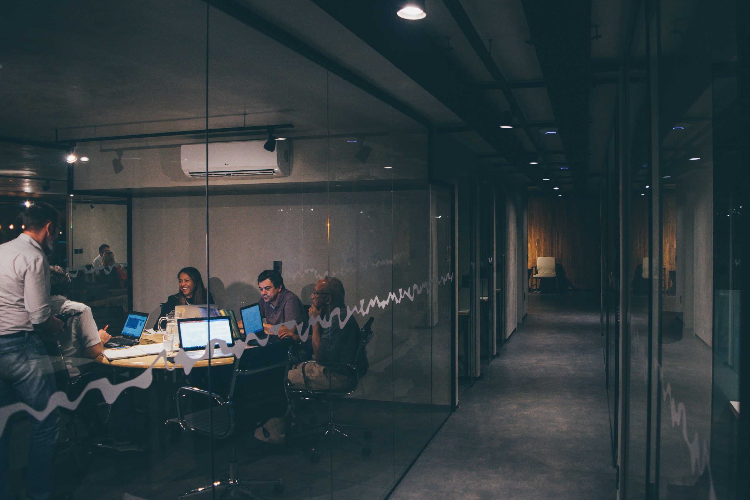 Photo of 5 young adults sitting at a conference table in a glass-encased room. They are talking and working on their laptops.