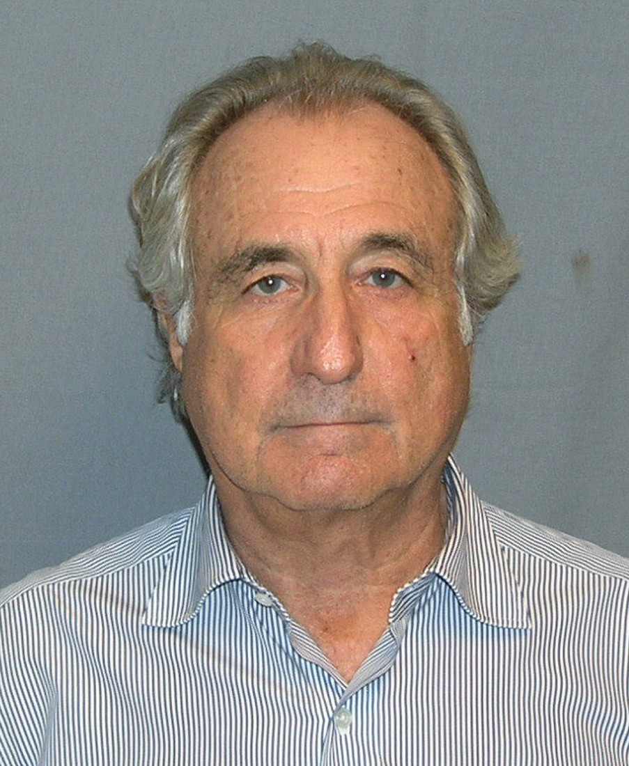 A photograph of Bernie Madoff standing in front of a blue background. He's an older white man with white hair and a flat smile. He's wearing a striped collared shirt.