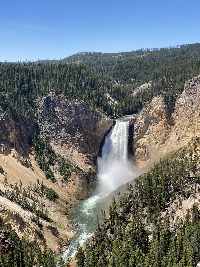 Large waterfall flowing into rushing river running through a steep-sided valley, with yellowish rocks and trees covering the valley slopes.