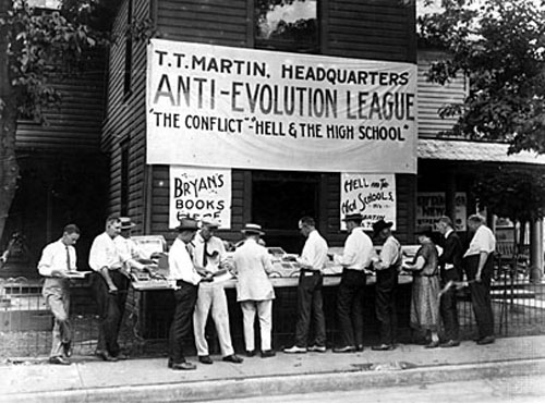 A group of 12 people stand below a banner that says "T.T. Martin Headquarters Anti-Evolution League 'The Conflict' - "Hell & The High School'