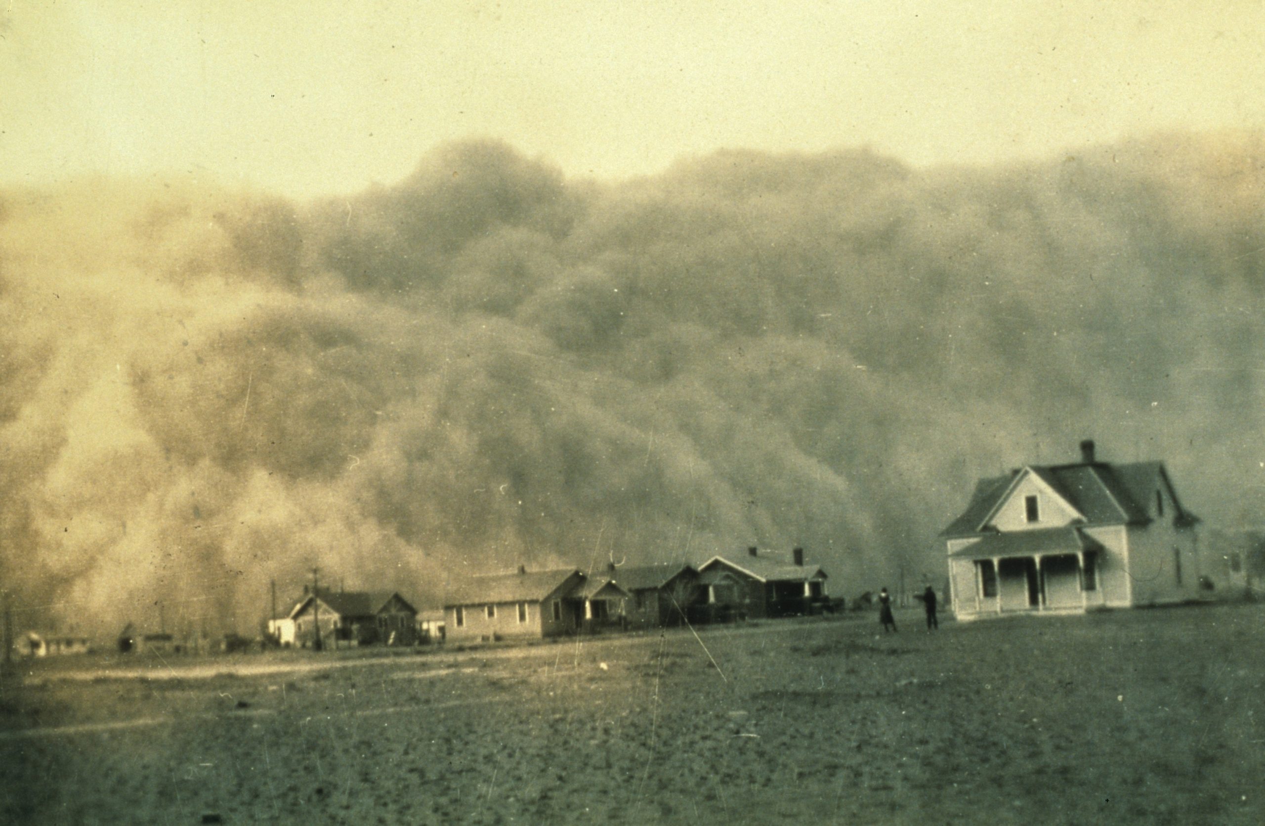 Black and white photo of a few houses on flat land along with two people standing near a house; a giant wall of dust is seen approaching in the background of the photo.