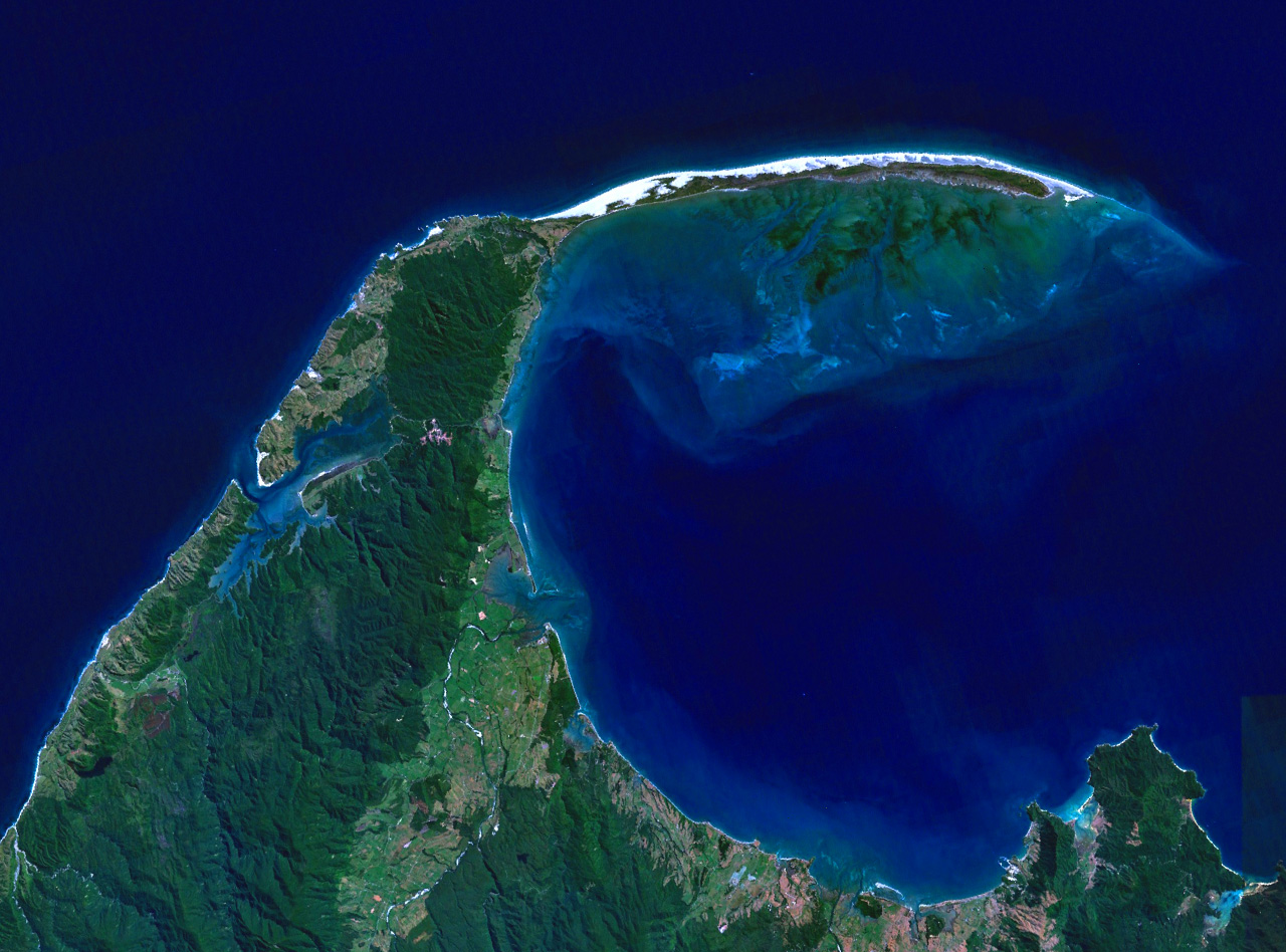 Satellite image of a green crescent-shaped landform with the tip of the crescent formed from sand extending into the ocean.