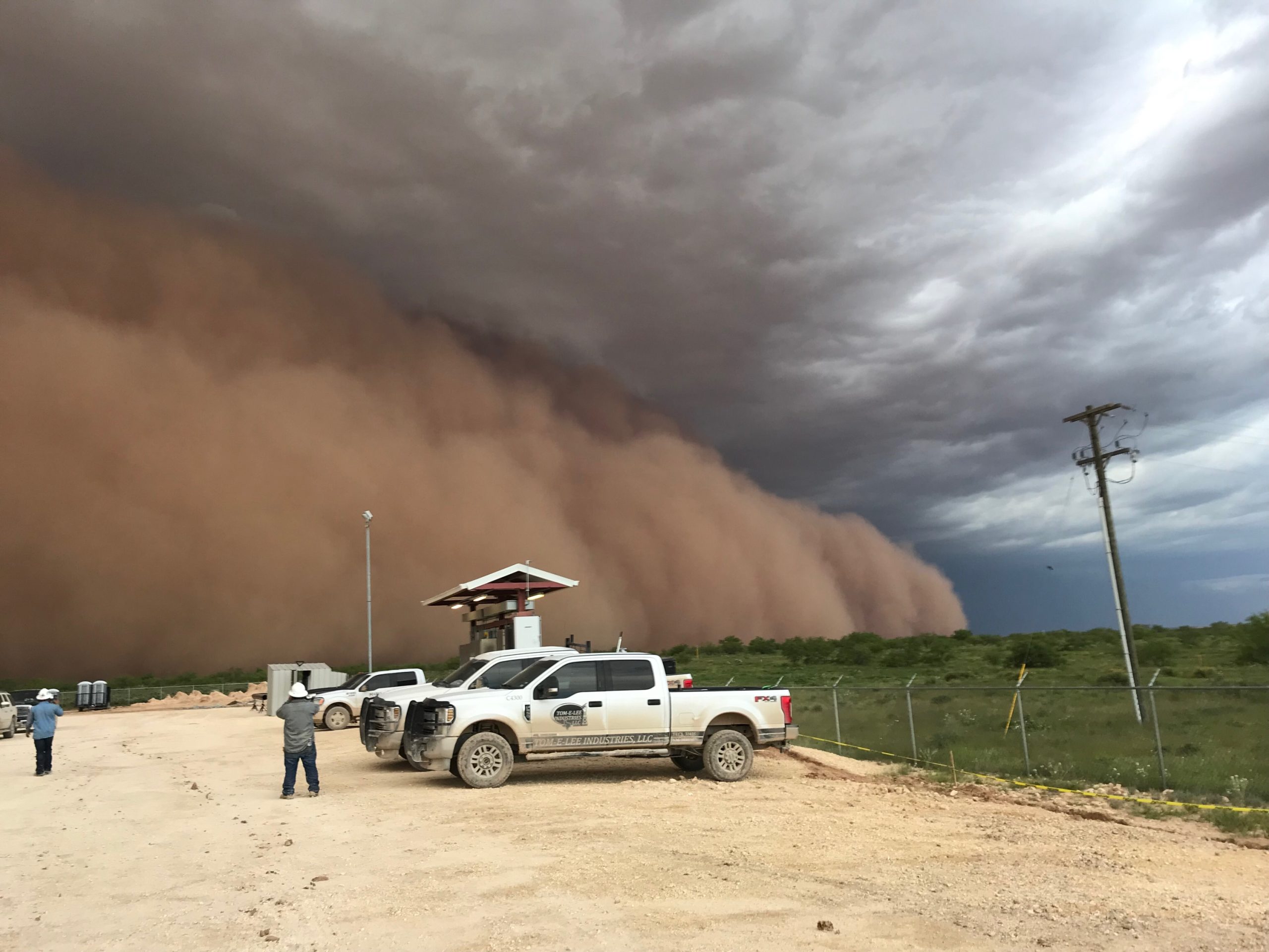 A few trucks on dry, flat land. From ground to sky is a dark brown dust cloud that you cannot see through.