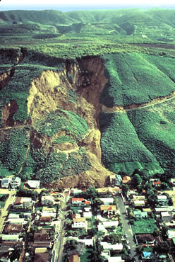 Vegetated hillside with a large section that has slumped down onto several houses in a neighborhood at the base of the hillside.