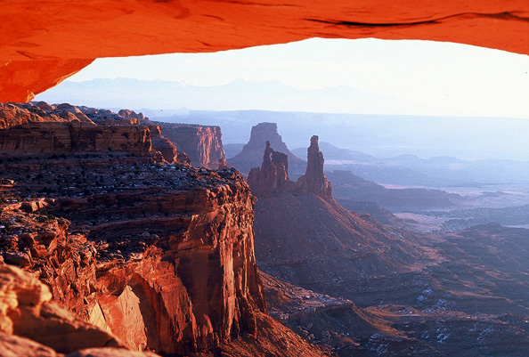 Photo through a sandstone arch of a desert landscape with steep sandstone cliffs, distant spires, and a flat basin.