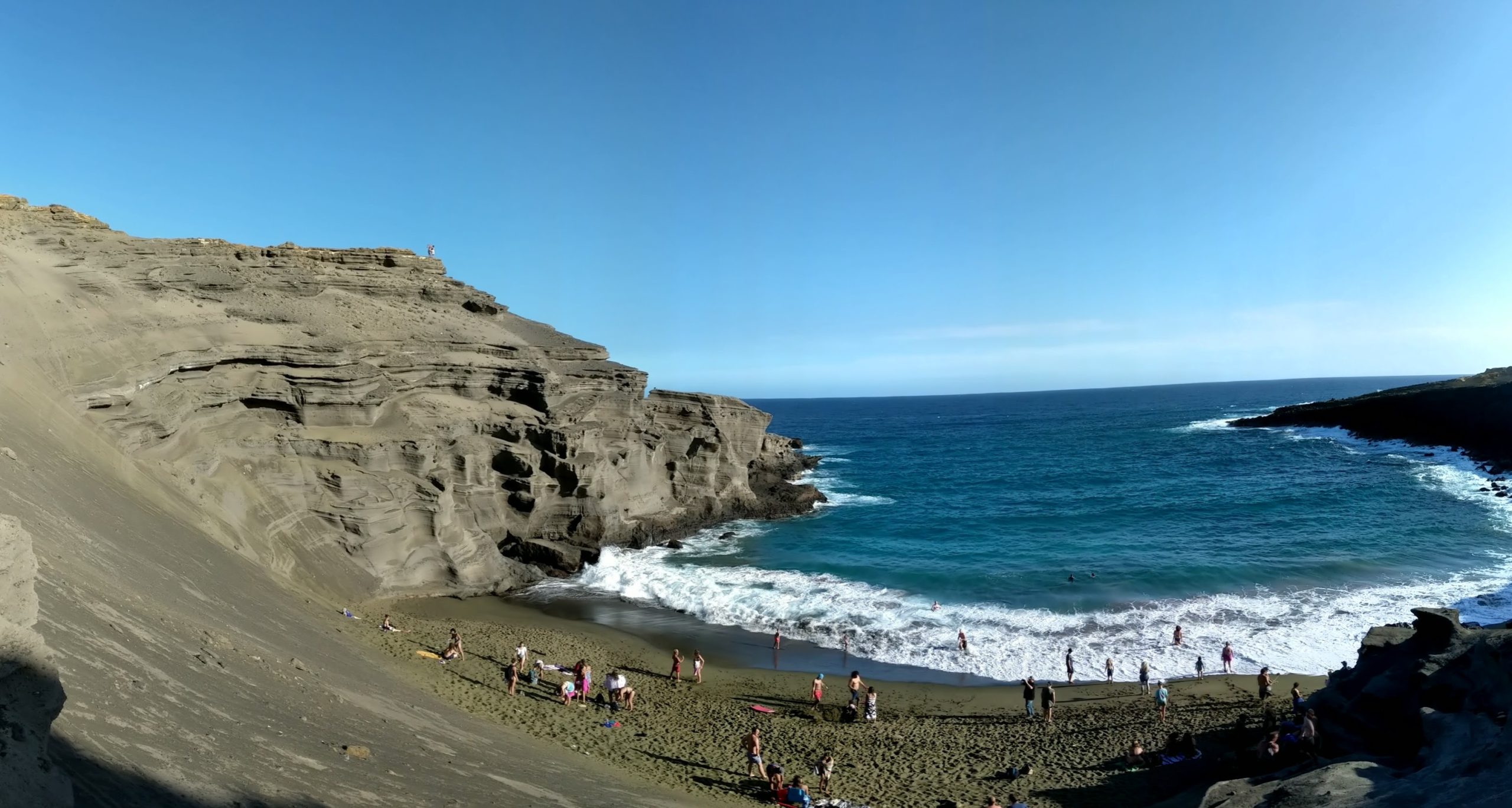 Beach cove with green sand and numerous people; surrounding the cove are black layers of volcanic rock.