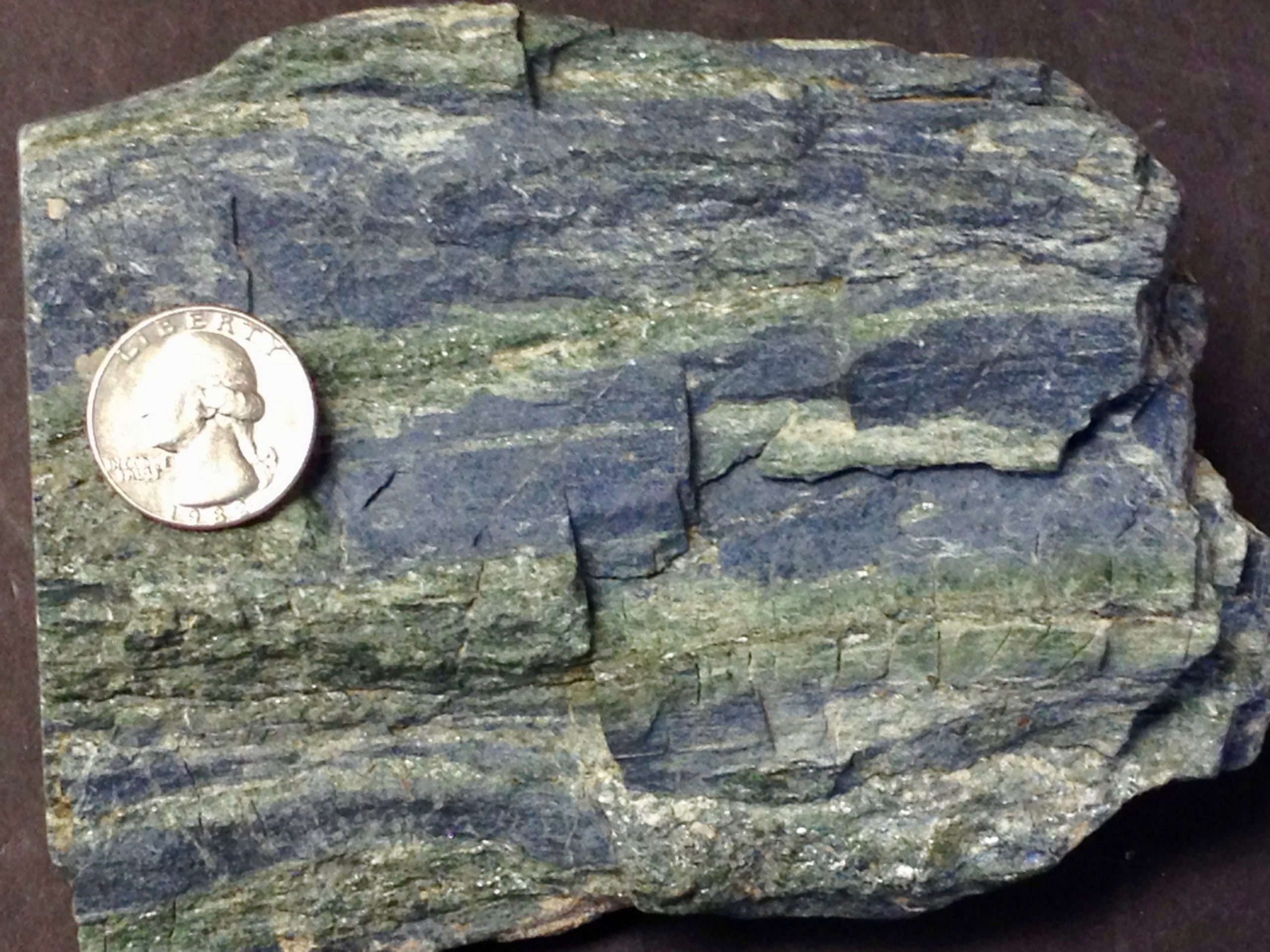 Sample of dark blue rock with dark green bands throughout; silvery mica grains can also be seen sparsely throughout the sample; a US quarter rests on the sample for scale.