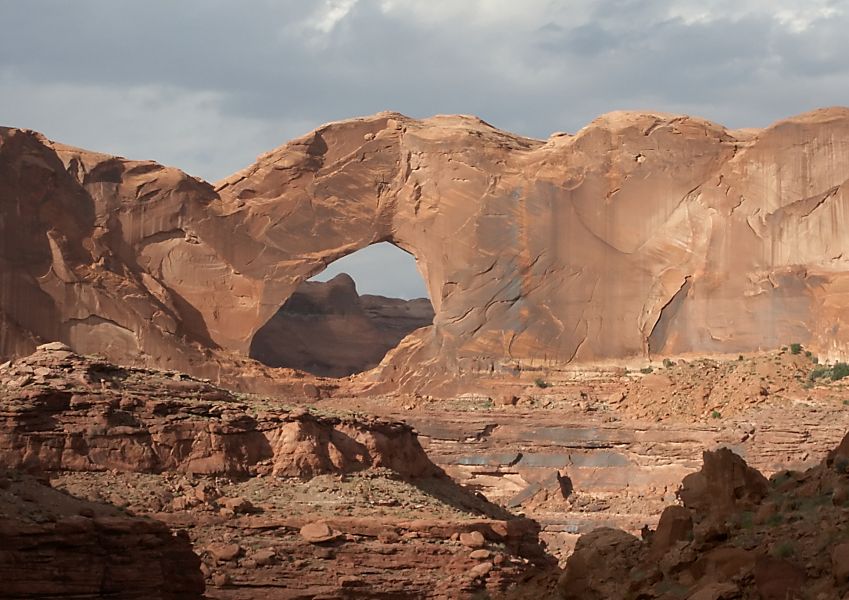Landscape view of thin layers of tan and brown sedimentary rocks with a thick layer of tan to brown rock on top, featuring a large hole in the rock that forms an arch.