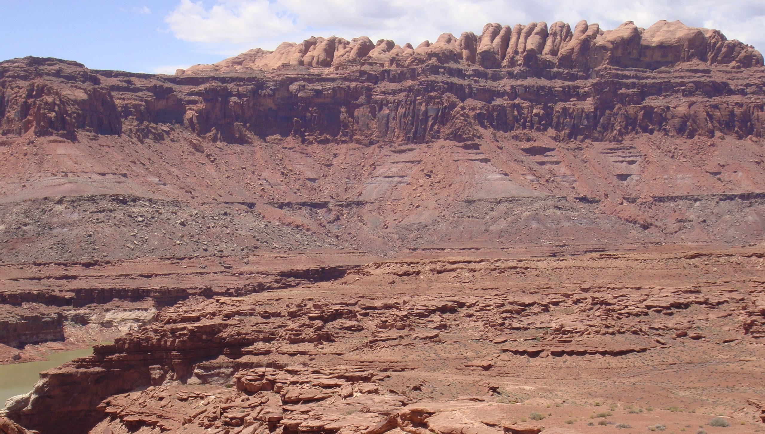 Landscape of flat layered reddish brown rocks in the foreground; in the background are slopes of gray and red rock fragments leading up to vertical reddish brown and tan cliffs above, consisting of flat-lying sedimentary rock layers.
