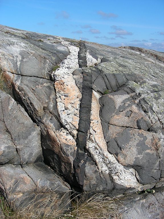 Rock outcrop with a nearly vertical white-tan igneous rock cutting through a larger, gray domed rock; there's a second thin black vertical igneous intrusion cutting through both the white intrusion and gray domed rock.