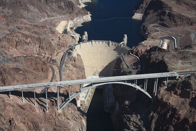 Aerial view of a large dam. The water level is much higher behind the dam than in front of it, and a road bridge runs across the canyon in front of the dam.