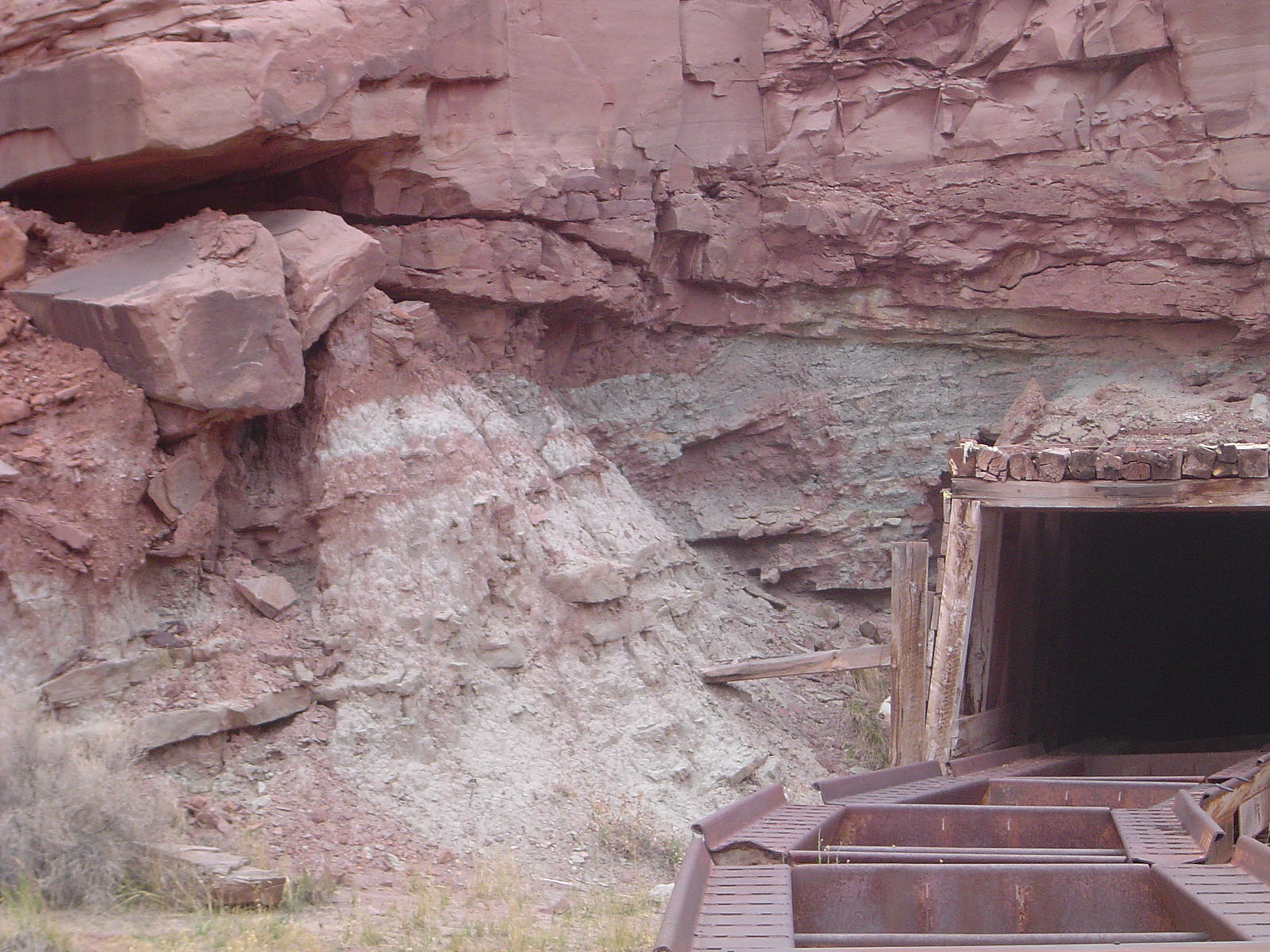 Dark mining shaft with rusted cart tracks leading into it. The shaft is carved into a cliffside composed of alternating red and whitish green sandstone and mudstone.