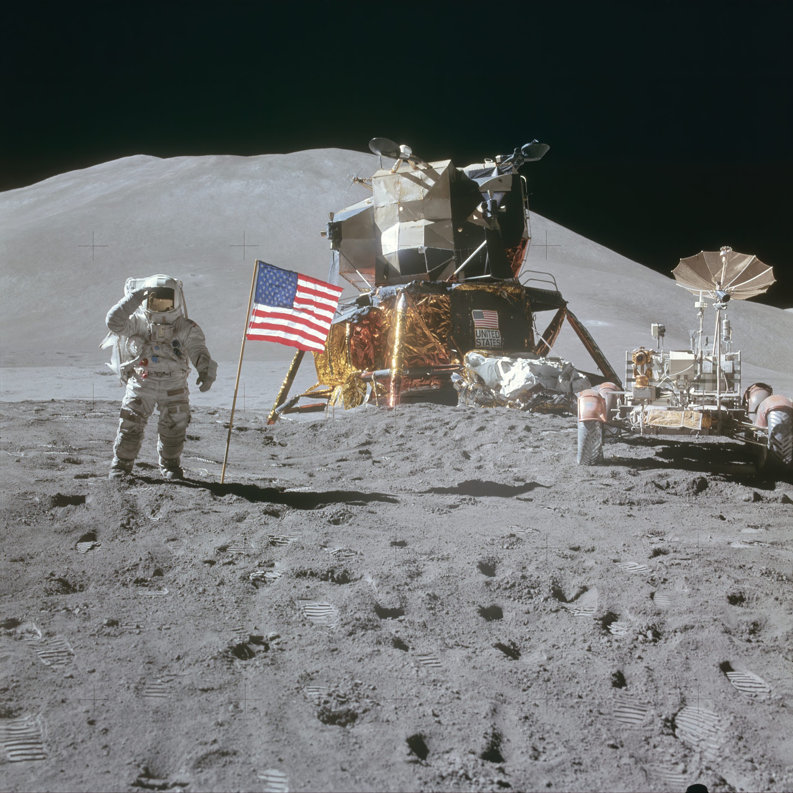 Photograph of Astronauts on the Moon. At center is the landing module, and to the right is the Lunar rover used by the Astronauts to travel large distances from the landing site. At left an Astronaut salutes the American flag placed near the lander. Scattered throughout the foreground are footprints in the Lunar soil.