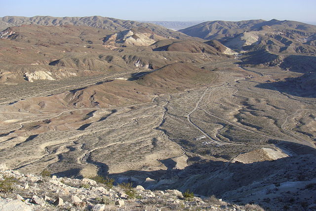 Landscape with a broad, tan valley dotted with low scrubby vegetation; a river from distant ridges splits and runs in numerous branches down the broad valley.