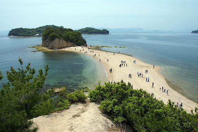 People walking along an elongate sand deposit that extends from the mainland out to a large vegetation-covered rock in the ocean.