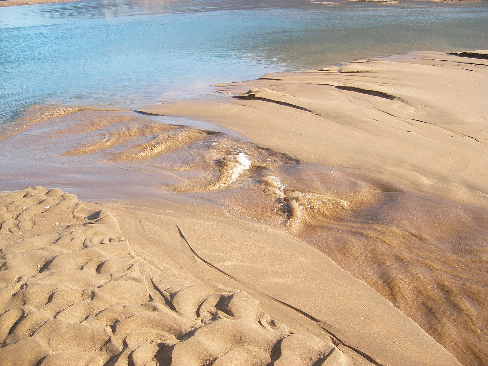 A shallow stream of water flows over a sandy bank toward a larger pool; the water stream has small waves in it as it travels over depressions in the sand.