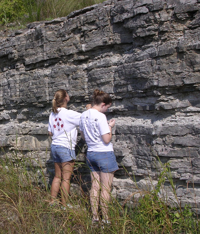 Two students are looking at a cliff composed of layers of gray rock.