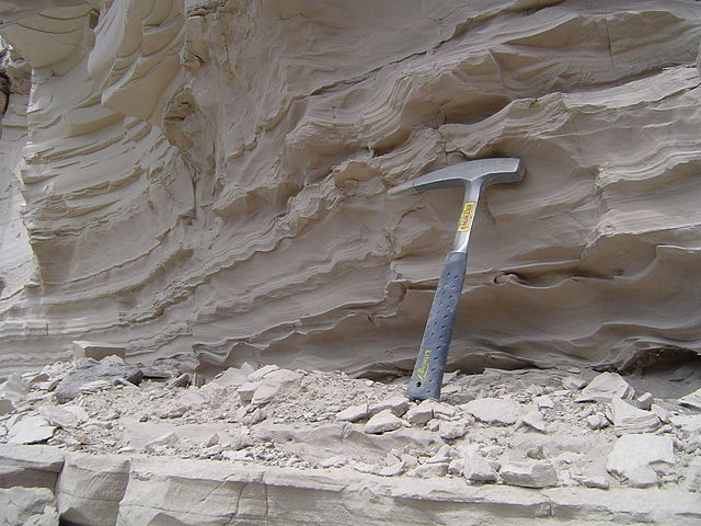 Light tan to gray chalky-looking rock cliff face with extremely thin laminations visible; a rock hammer leans against the outcrop for scale.