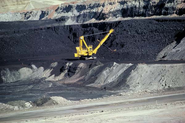 A large yellow machine is removing black coal from a terraced slope.