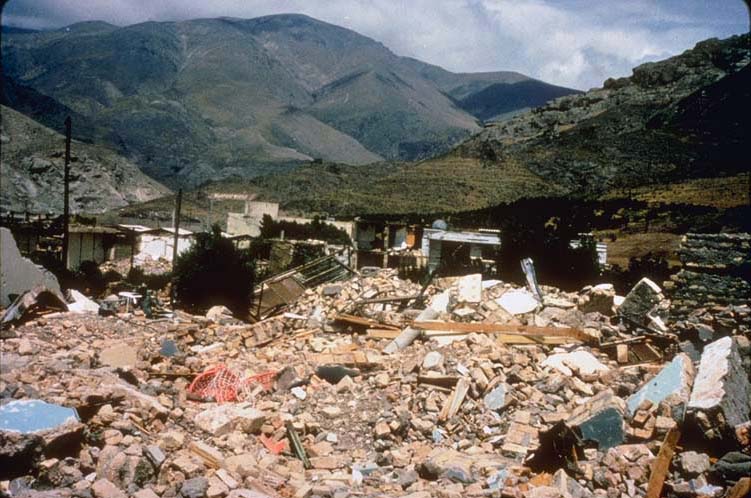 Color photograph of rubble from the remains of a collapsed building; in the background are vegetation-covered mountains.