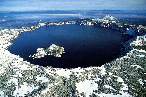 Aerial view of the top of a large mountain that has a large hole at its top, filled with deep blue lake water. There is a small cone-shaped island in the lake visible in the lower right of the photo.