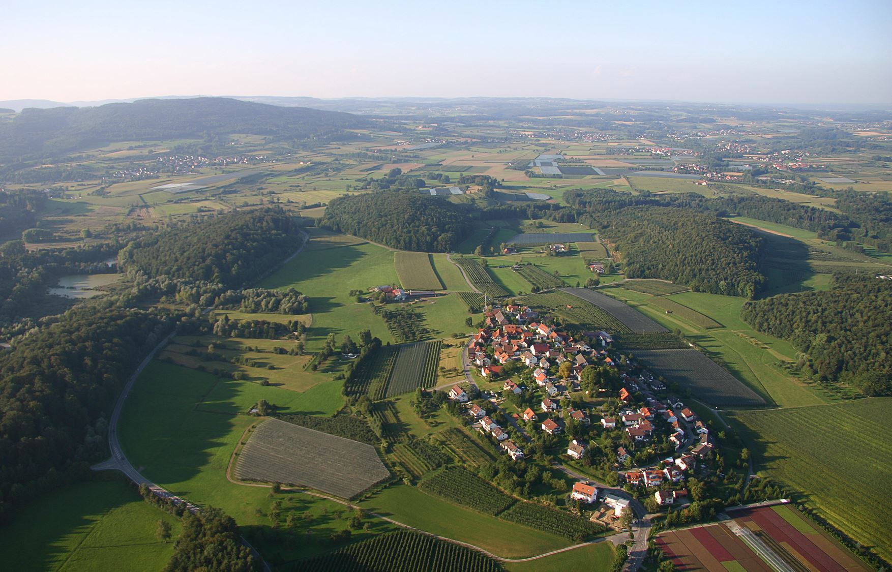 Aerial photograph of a small town surrounded by green landscape; numerous elongated asymmetrical teardrop-shaped hills are seen surrounding the town, which is also located on a small ridge.