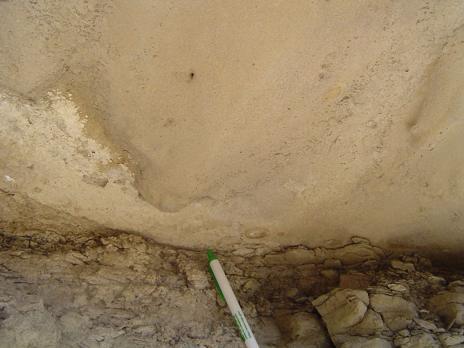 A view of the bottom of a tan rock layer with a bulge sticking out of the base toward the observer; a pen is next to the rock for scale.