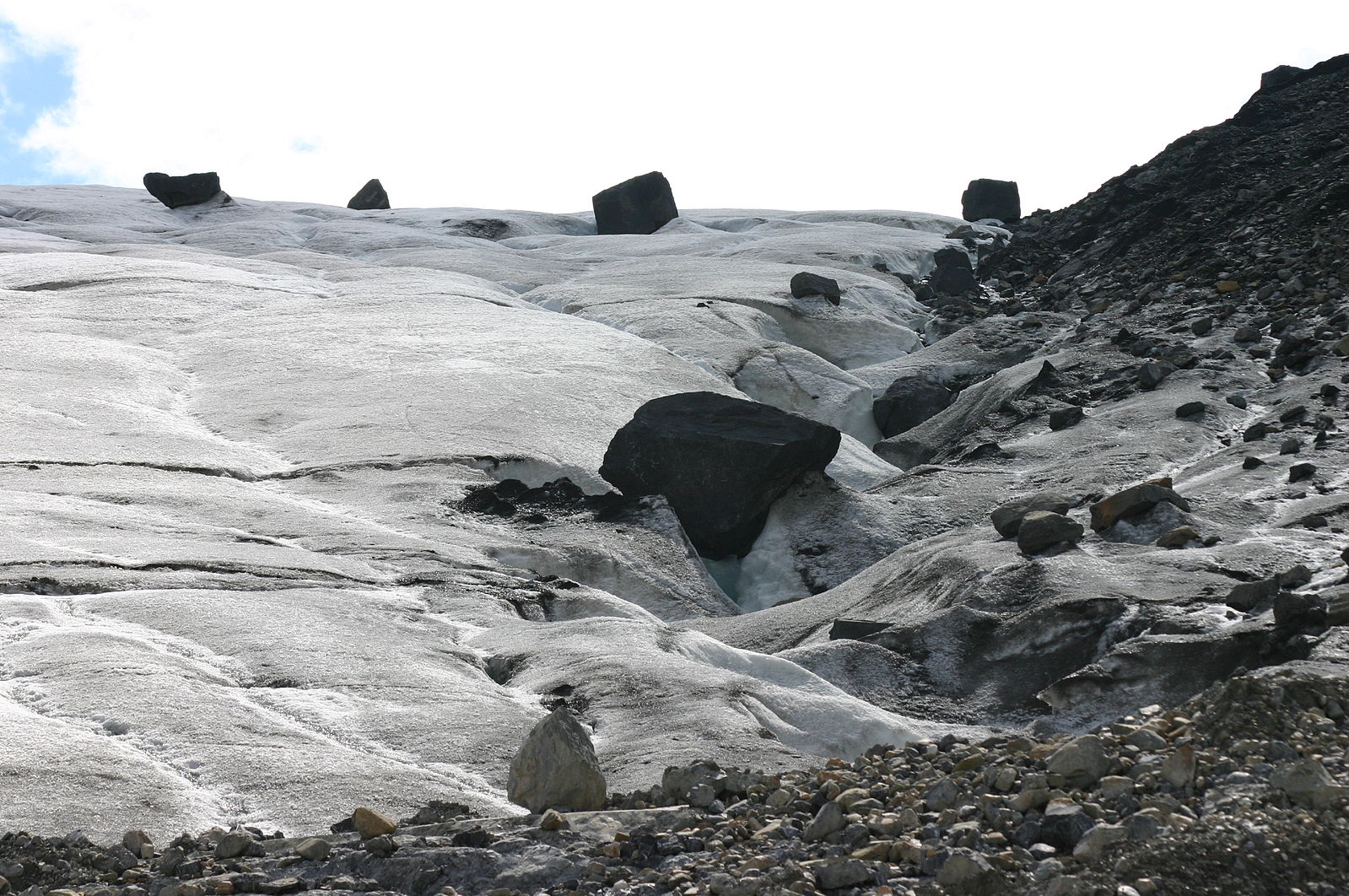 Along the edge of glacial ice, numerous broken pieces of tan, gray, and black rocks lay along the edge, ranging in size from large boulders to tiny bits.
