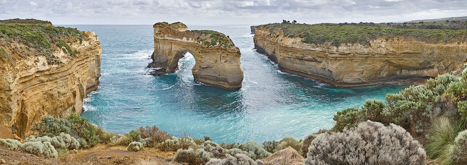 The arch is a rock formation in a cove of water with a hole in the middle which allows water to pass through; the surrounding landscape is composed of vertical tan cliffs.