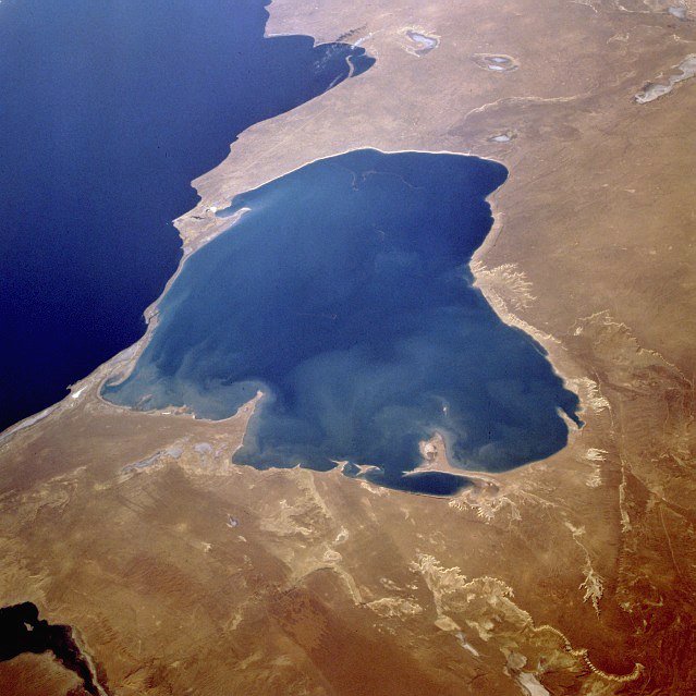 Satellite image of a brown, barren landscape with a large blue rounded body of water on the land; to the left of that body of water is a deeper blue larger water body, the Caspian Sea.