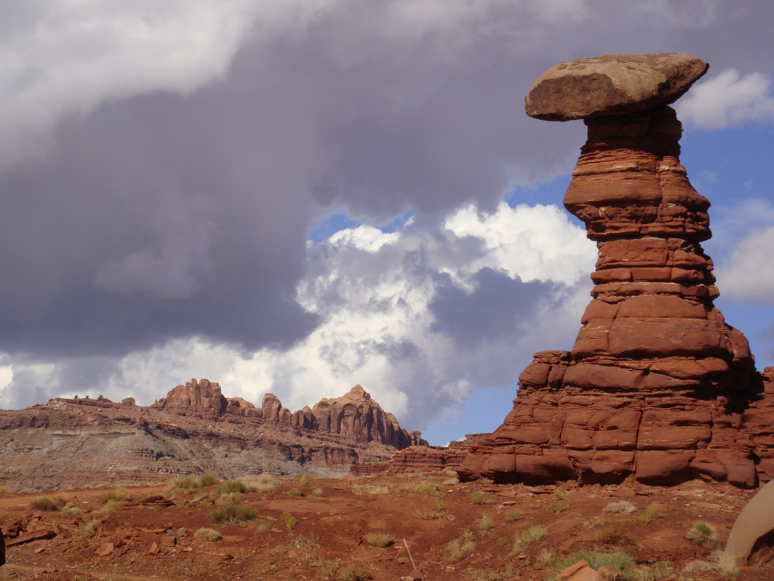 Tower of brick red rock with visible horizontal layers; the tower thins upward except for a larger, flat rock capping the top of the tower.