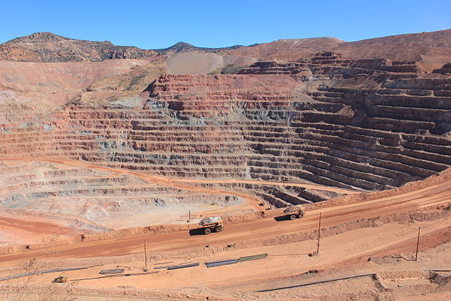 A large terraced open pit mine that contains both gray and red rocks; a haul truck is seen driving in the foreground.