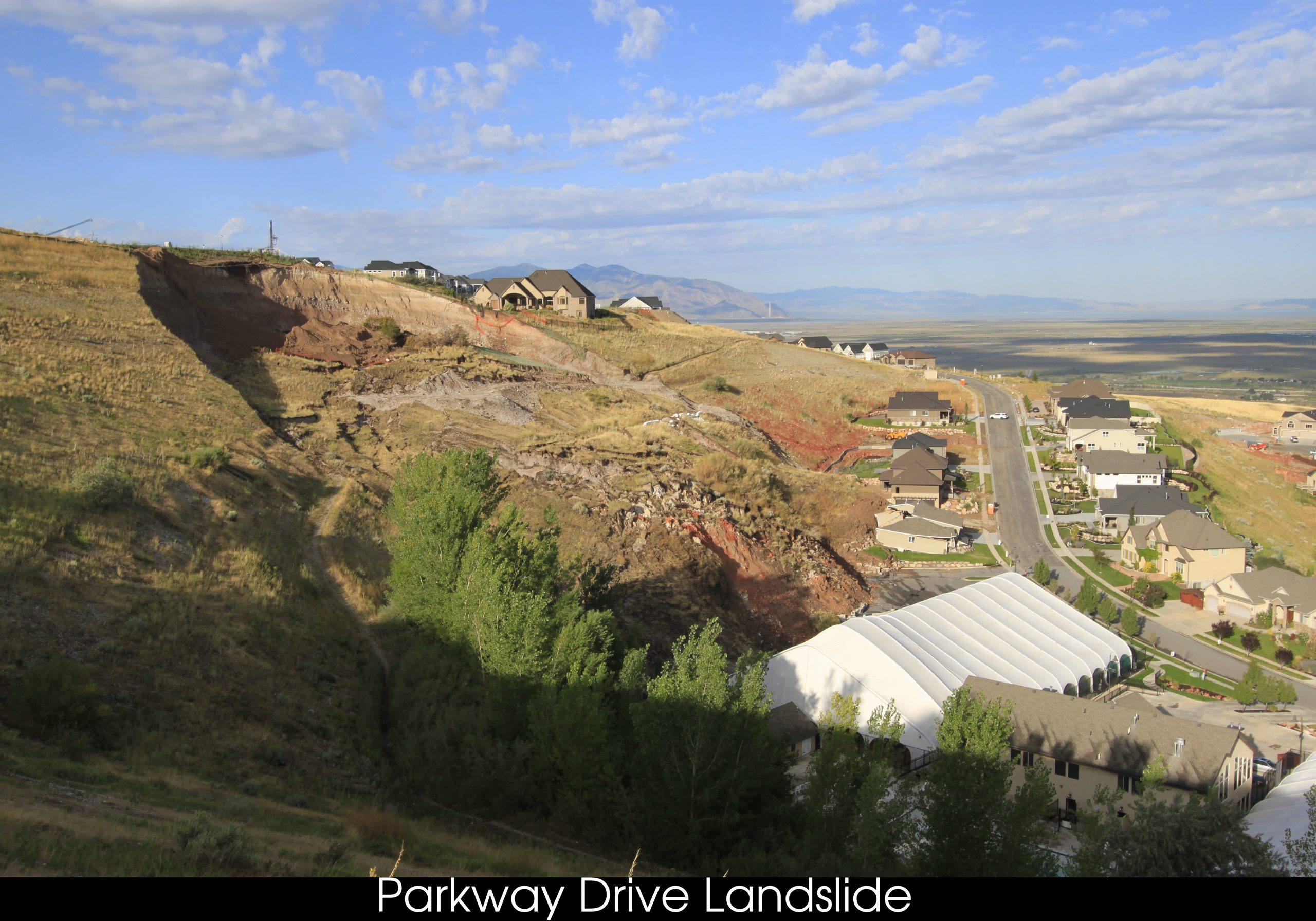 Vegetated hillside with a large slump of grass-lined material at the base of the hillside just above a row of houses; a tan sliver-shaped scarp can be seen in the hillside.