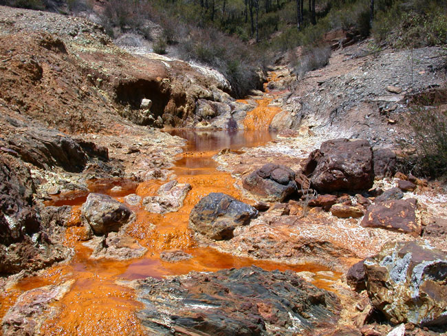 Bright orange river flowing toward the viewer. The land on either side of the river is rocky and barren.